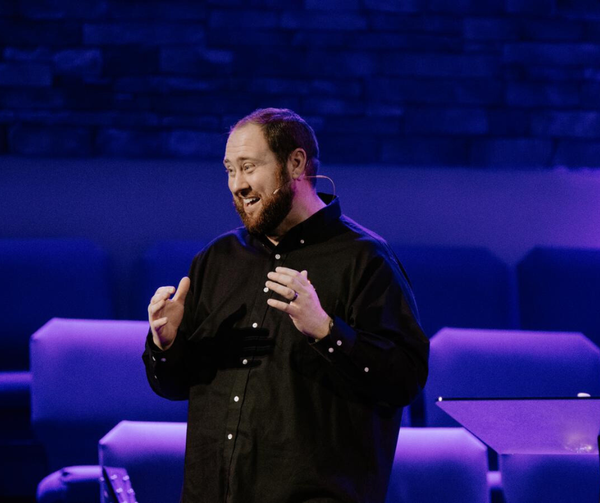 Man on a blue-lit stage speaking and gesturing, wearing a black shirt, with sofas in the background