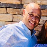 Smiling man in a light blue shirt posing outdoors against a stone wall