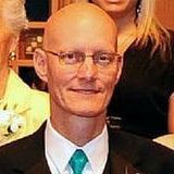 Bald man in glasses and suit, wearing a green tie, posing indoors with people in the background