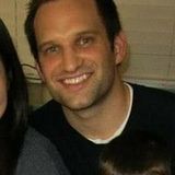 Smiling man in a black shirt indoors, seated beside another person.