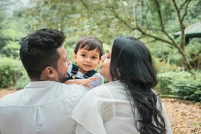 Family sitting outdoors in a park, holding a smiling toddler amid green trees