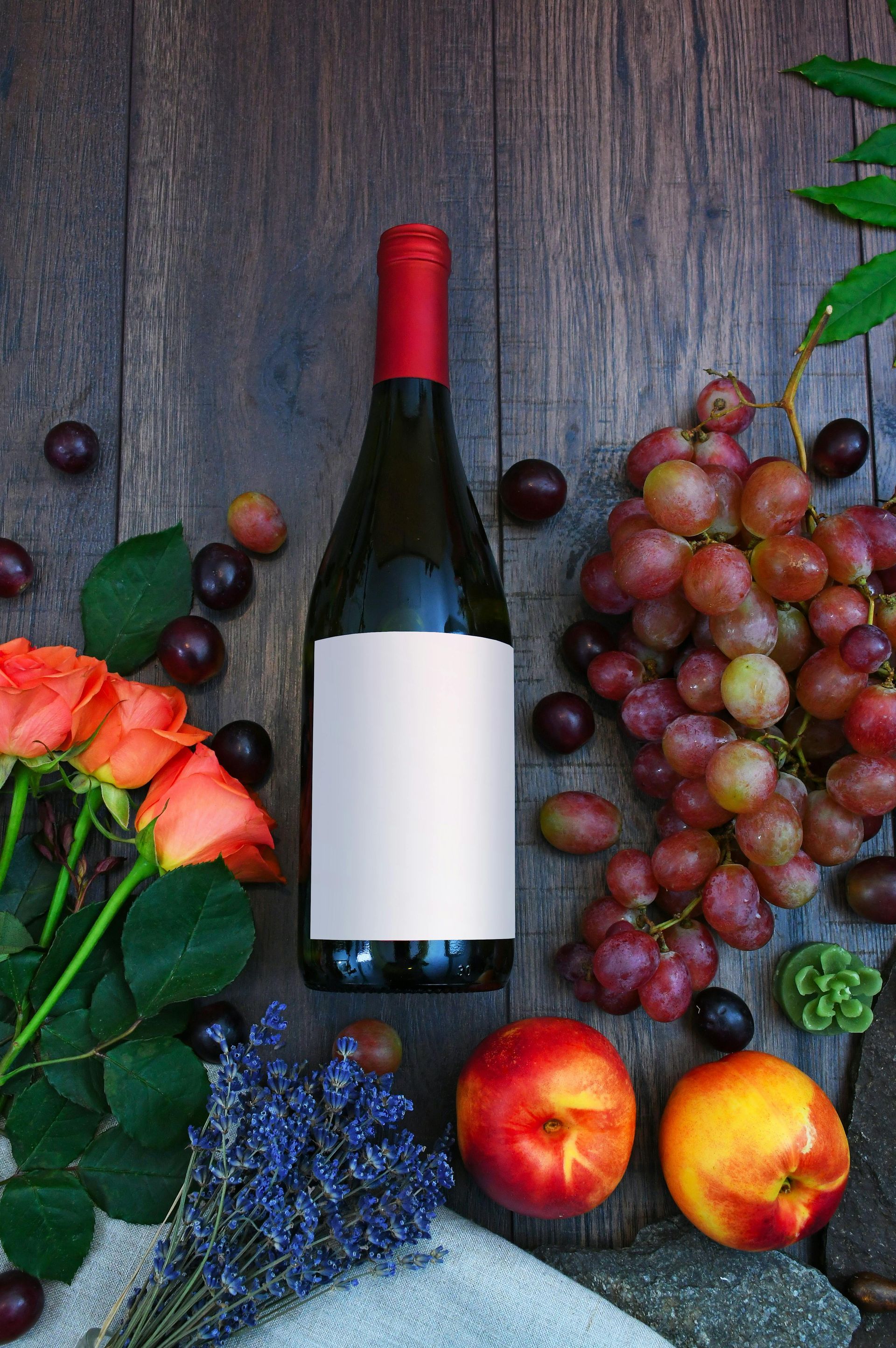 A bottle of wine surrounded by grapes , apples , roses and lavender on a wooden table.
