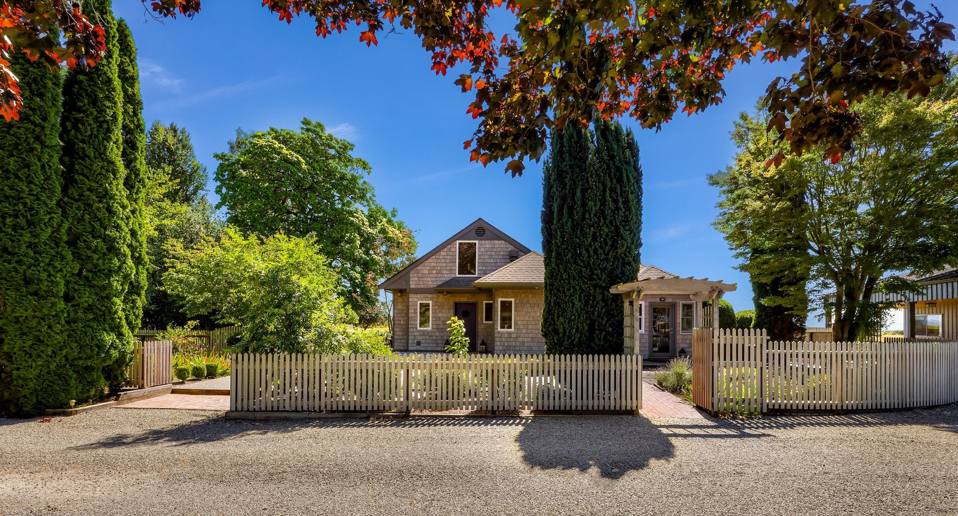 Charming shingle-sided home with a picket fence, mature landscaping, towering evergreens, brick pathways, and a garden patio framed by colorful fall foliage.
