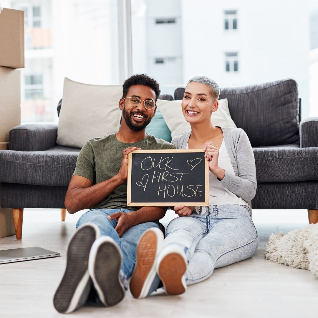 A man and a woman are sitting on the floor holding a sign that says our first house.
