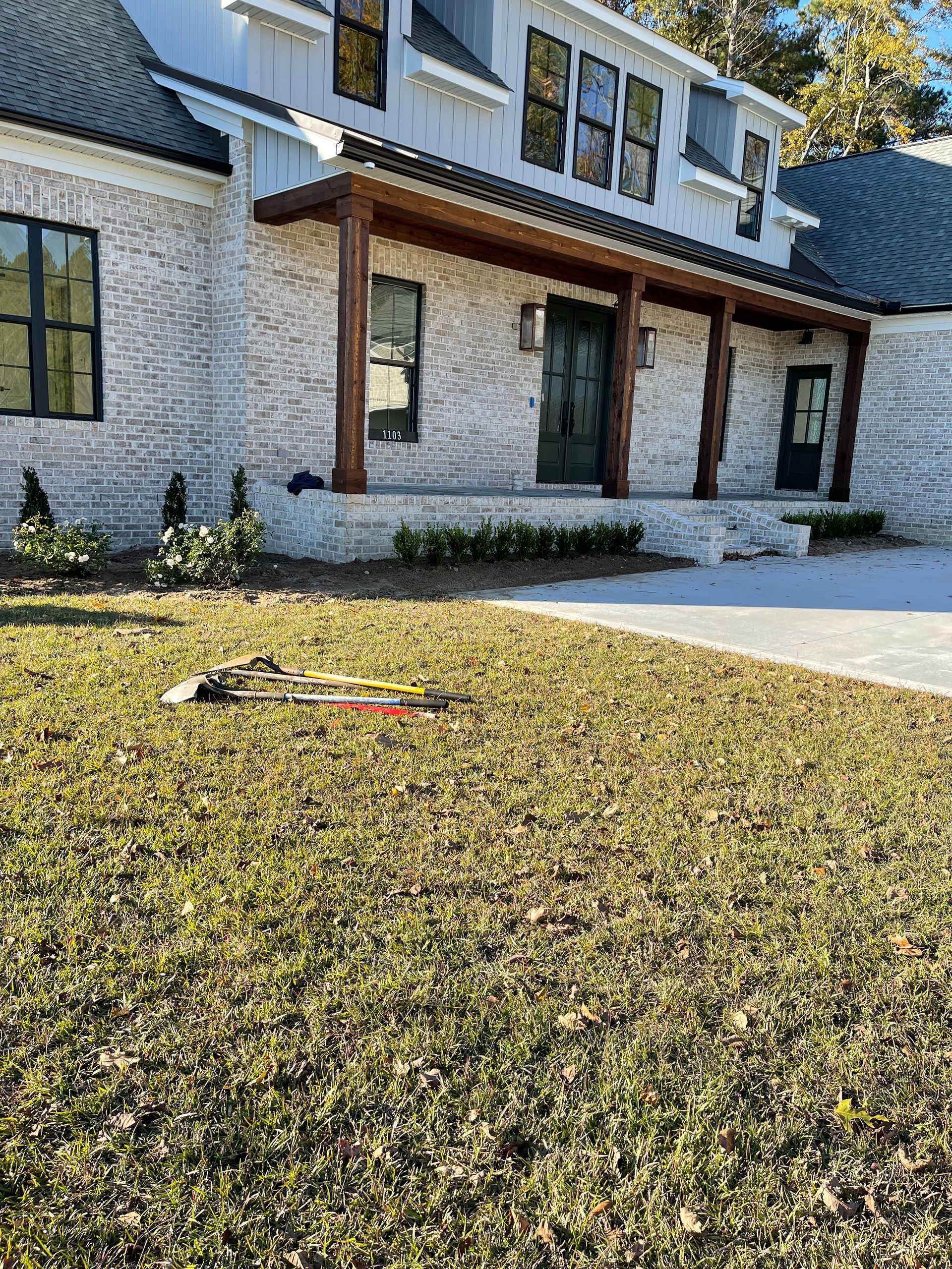House exterior with brick, dark trim, and a covered porch. Lawn in foreground, tools on the grass.