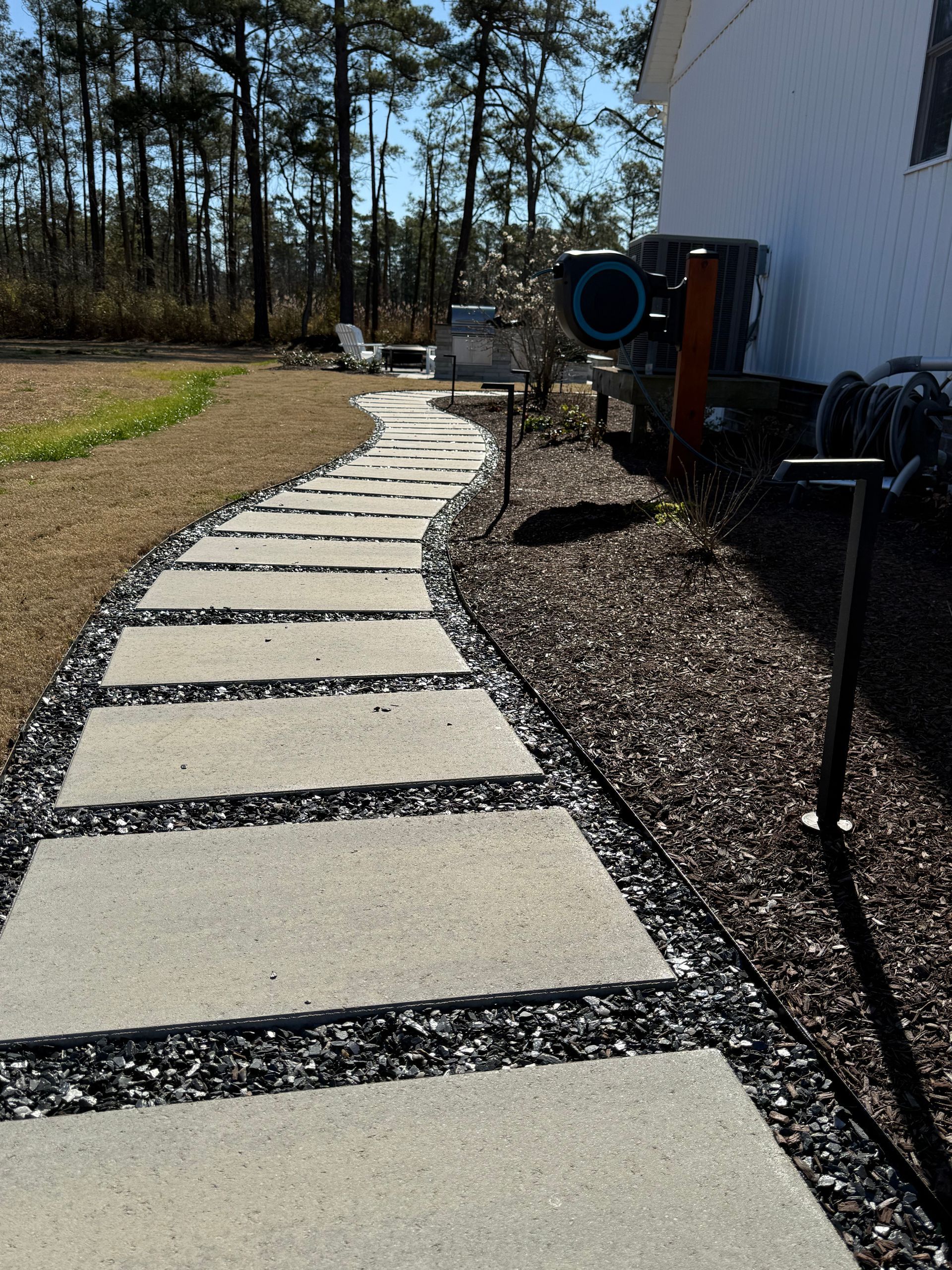 Concrete stepping stones lead through a yard with black gravel borders, near a white building.
