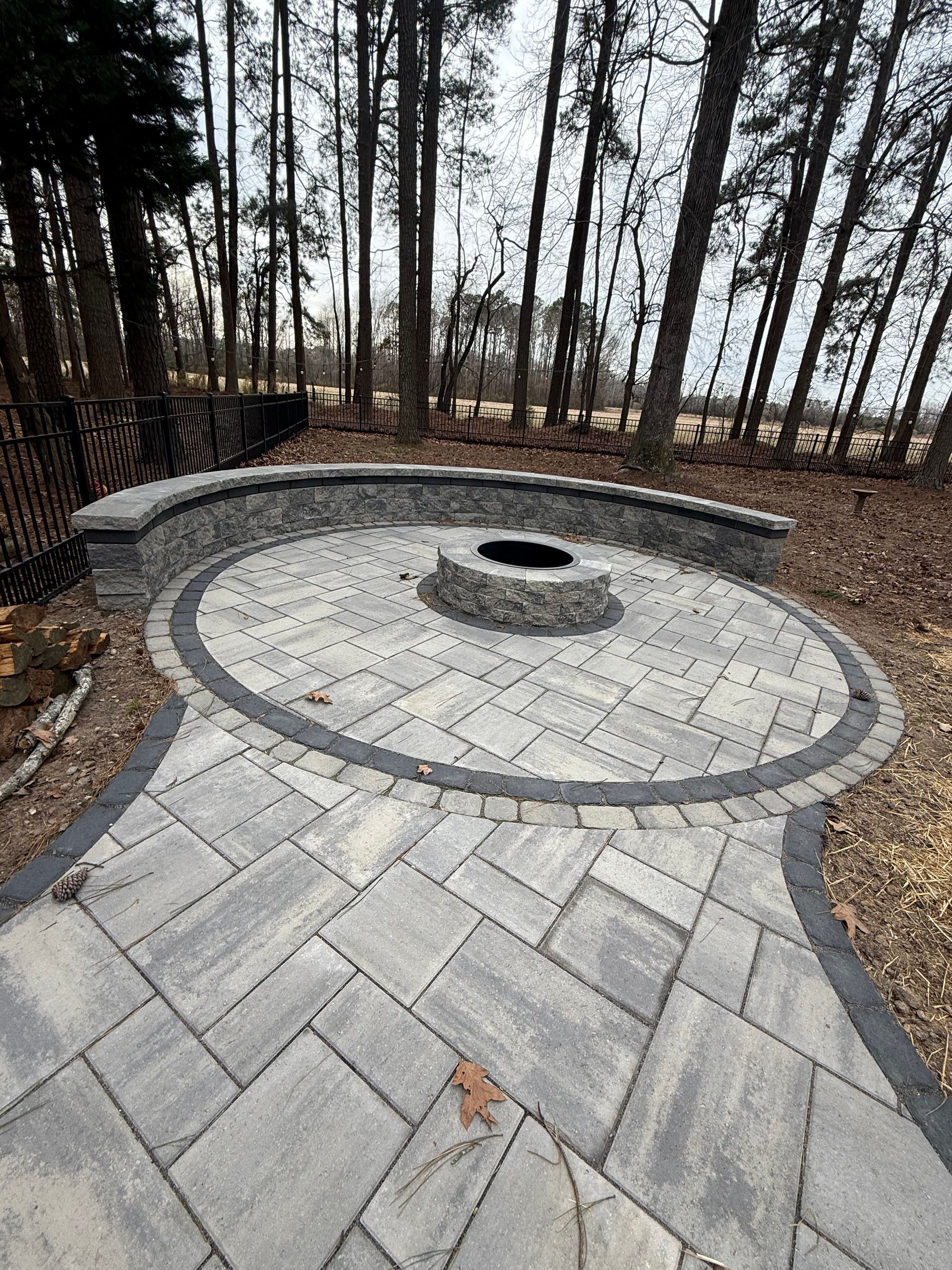 Stone patio with circular fire pit and curved seating wall, surrounded by trees.