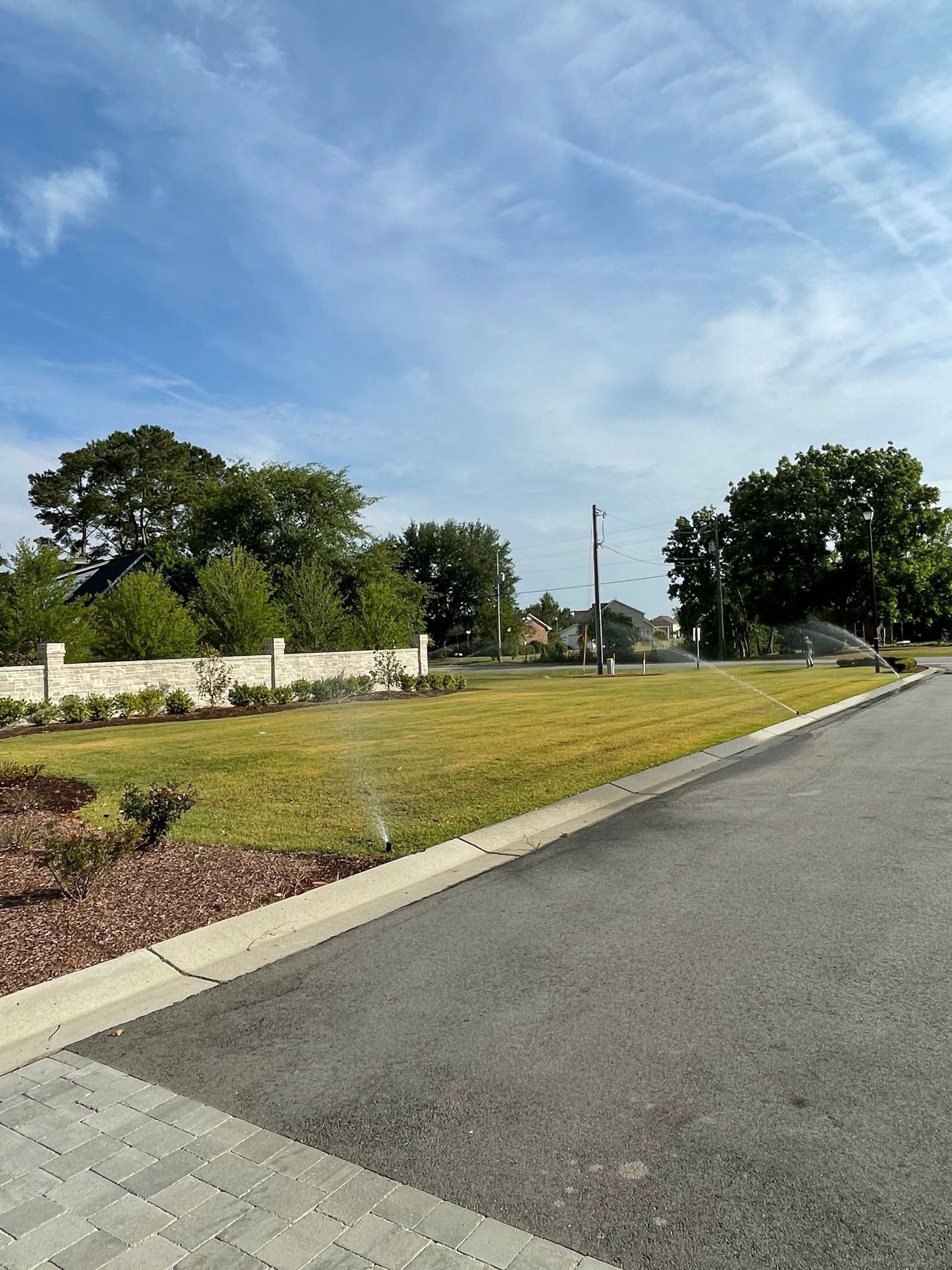 Lawn being watered, next to a paved road and brick wall. Trees and a blue sky in the background.