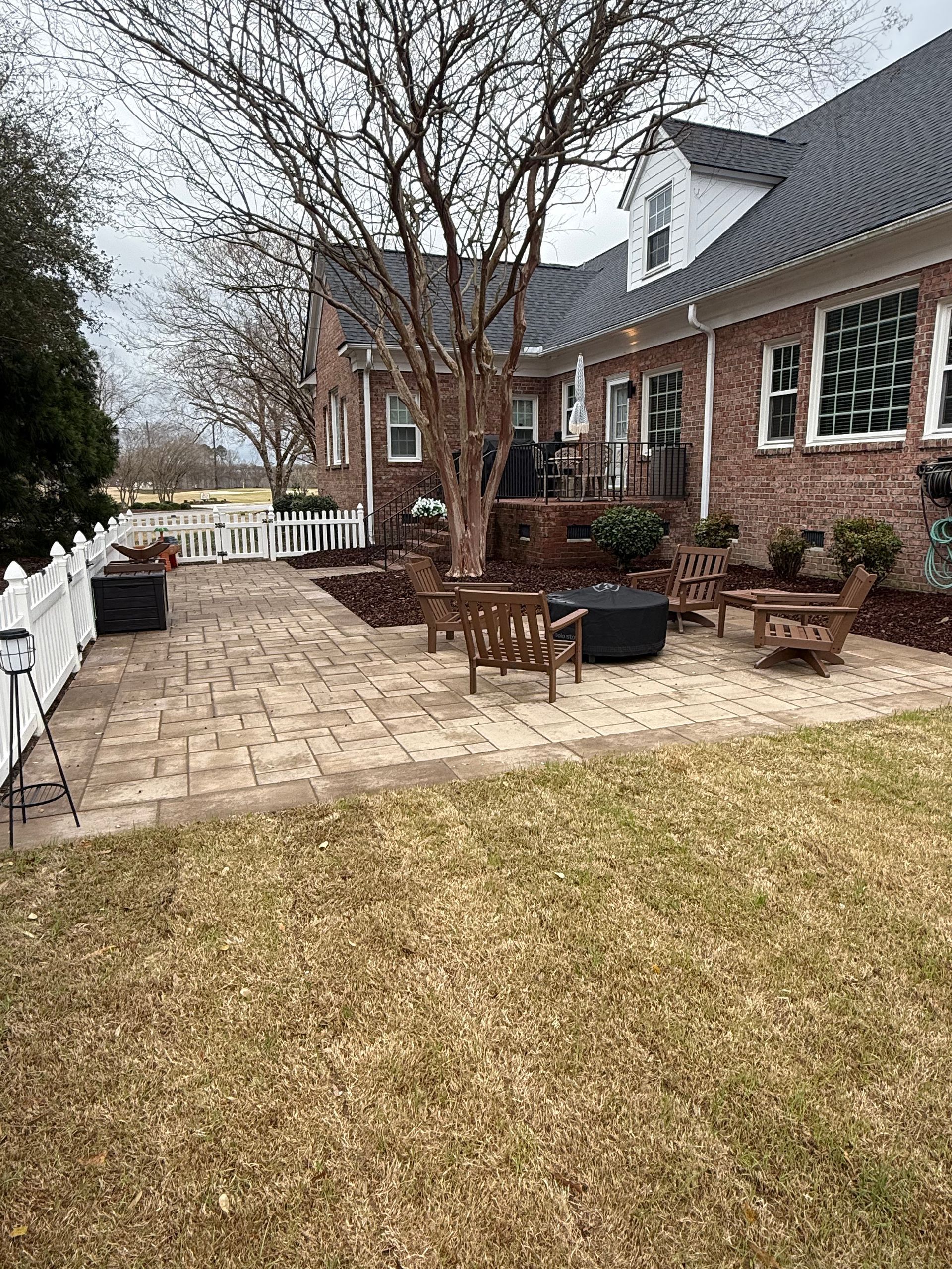 Brick patio with wooden furniture in front of a brick house with a white picket fence.