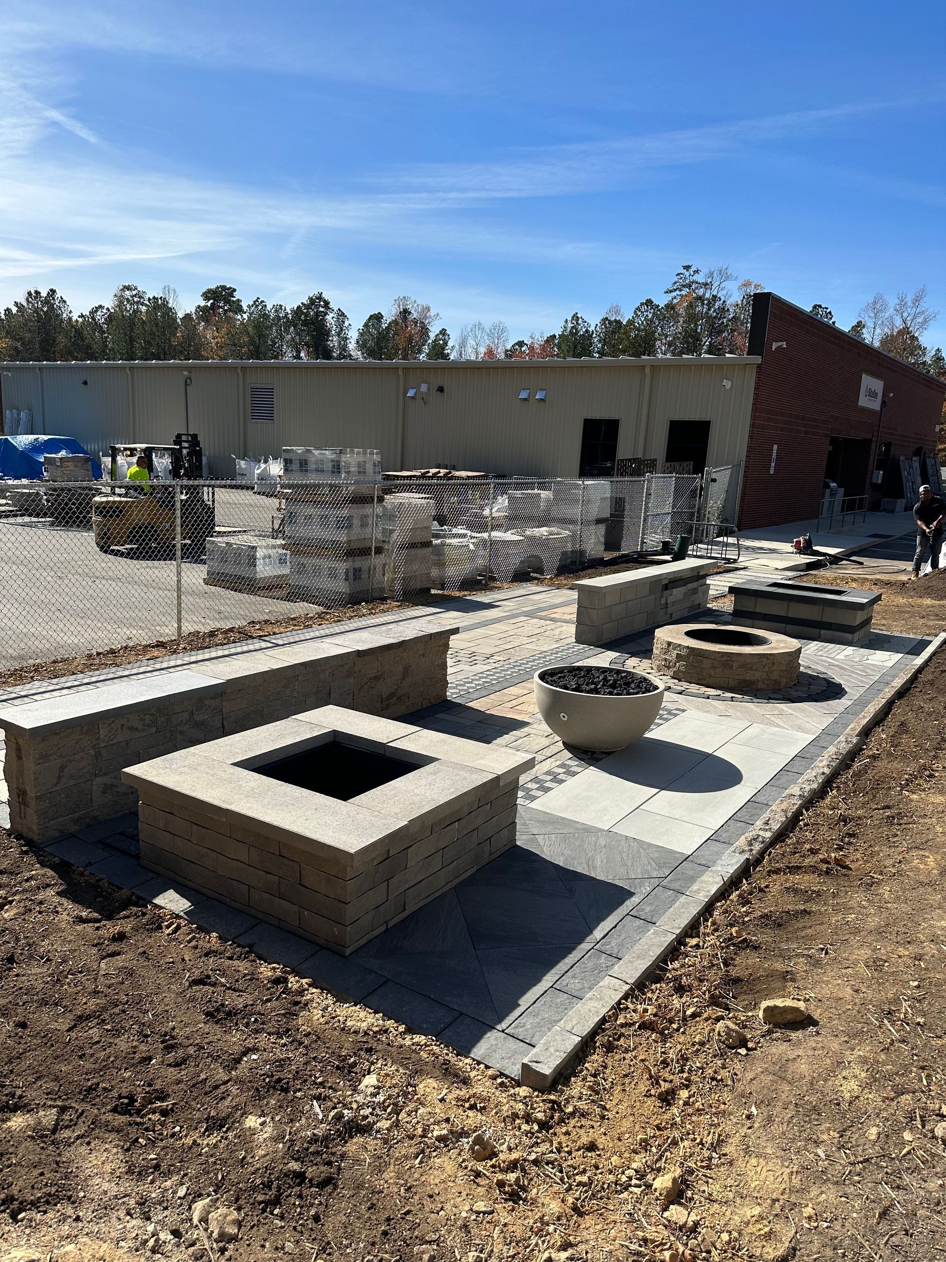 Outdoor patio under construction with fire pits, seating, and stacked pavers near a building.