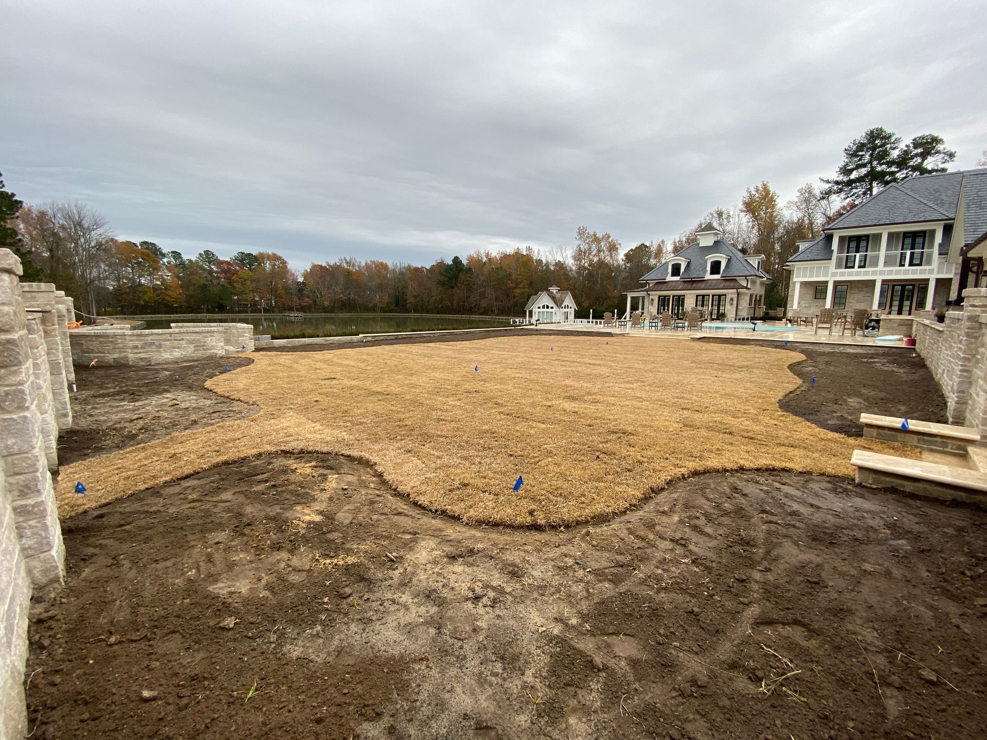 A large yard with straw covering, partially enclosed by stone walls and a house in the background under a cloudy sky.