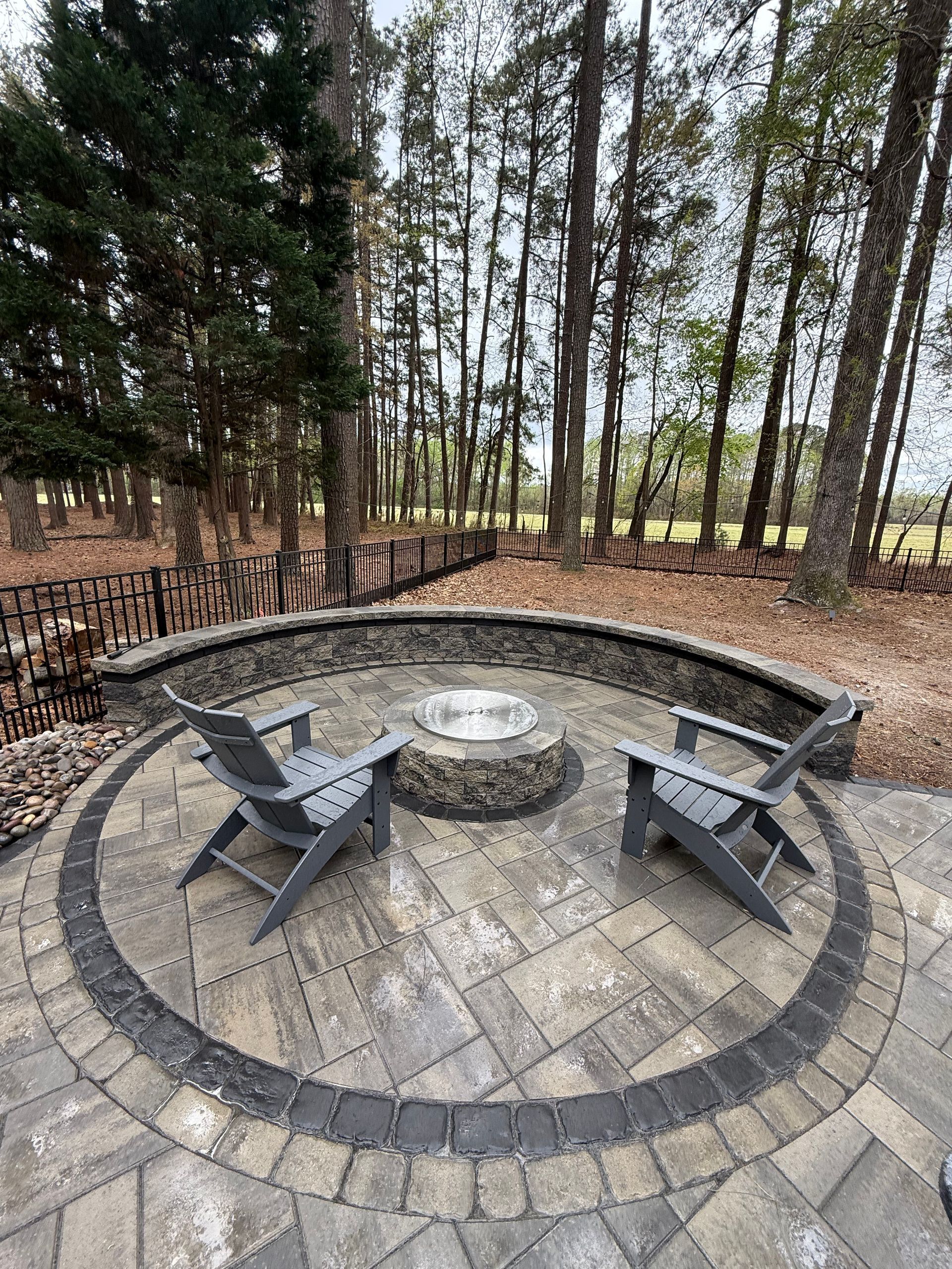 Two gray chairs face a stone fire pit on a circular stone patio. Dark stone border, trees in the background.