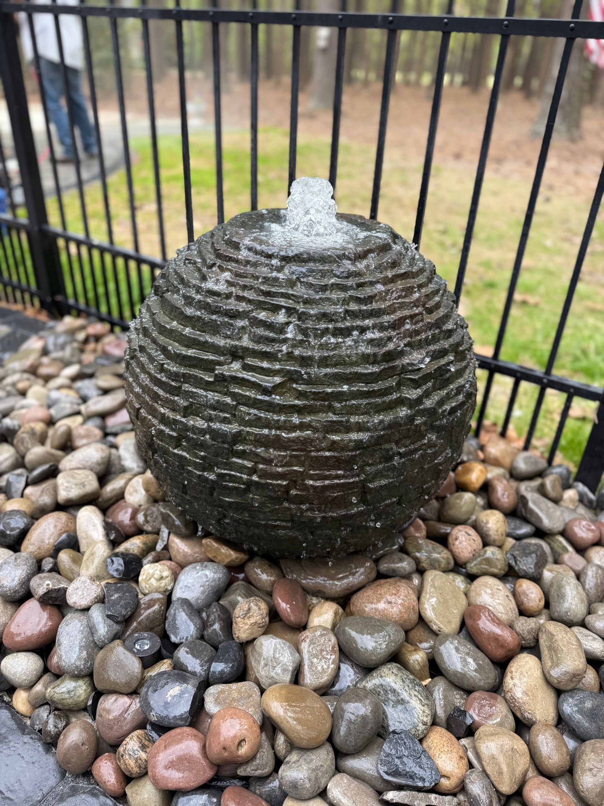 A water fountain in the shape of a textured sphere, surrounded by smooth, wet river stones.