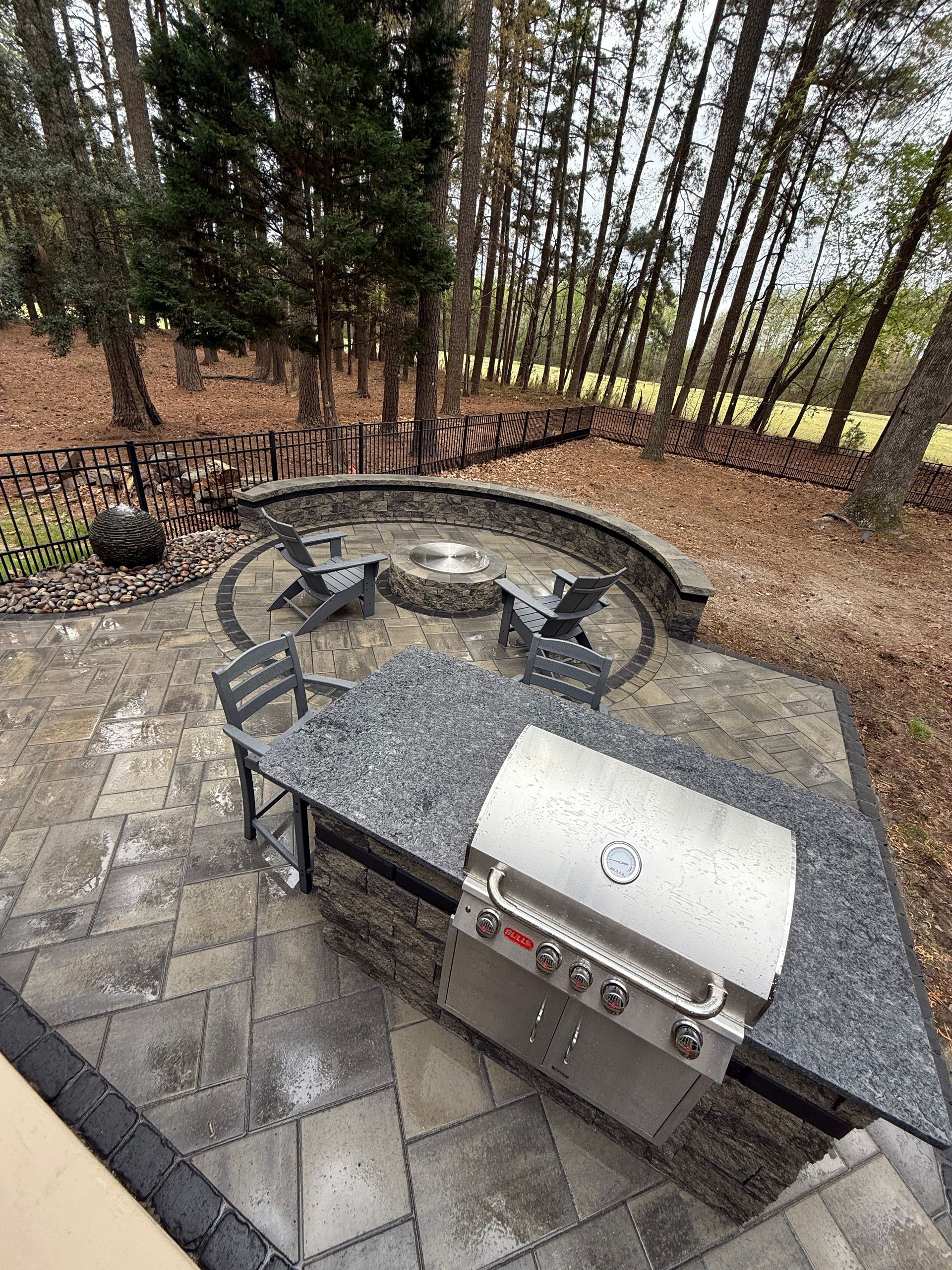 Outdoor patio with a grill, fire pit, seating, surrounded by trees.