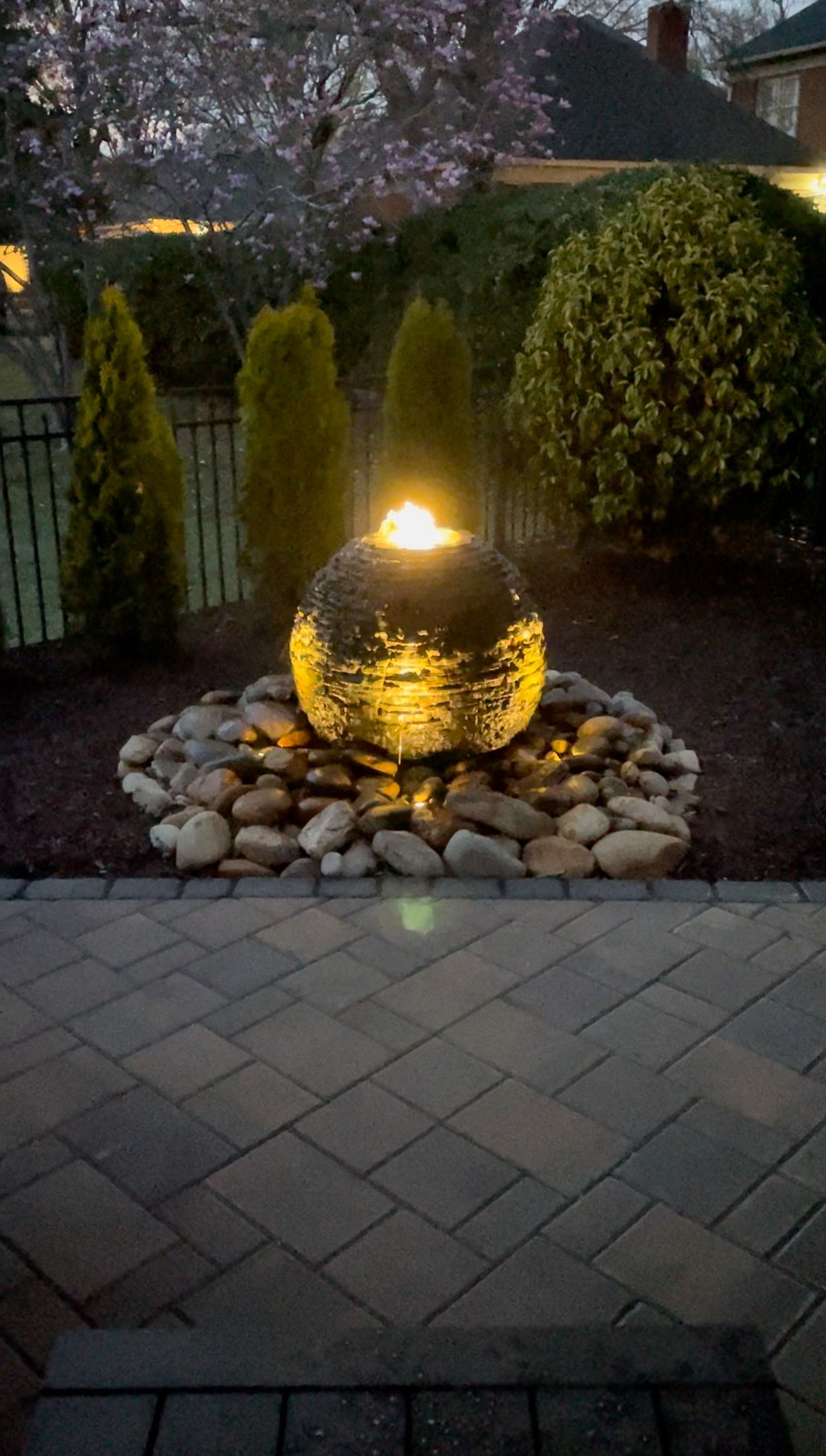 Glowing spherical water fountain in a garden, surrounded by rocks and evergreens.