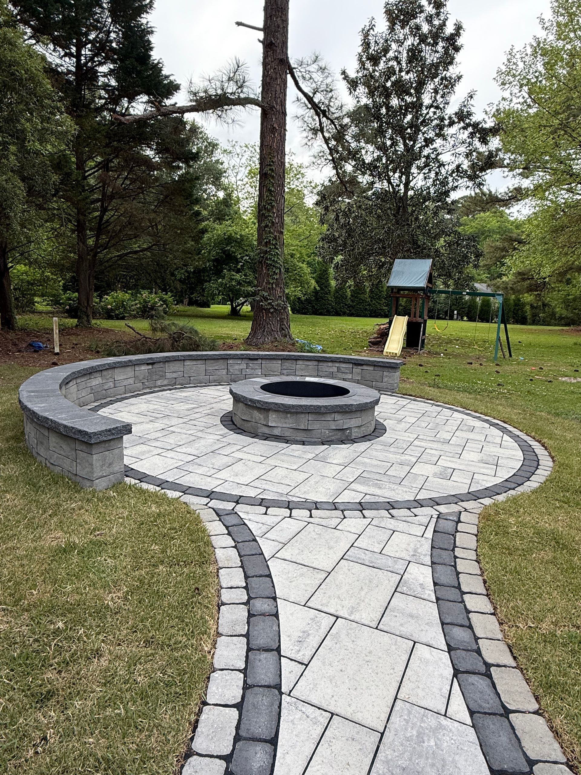 Stone patio with a fire pit, curved seating, and a pathway in a grassy backyard, near a playground.