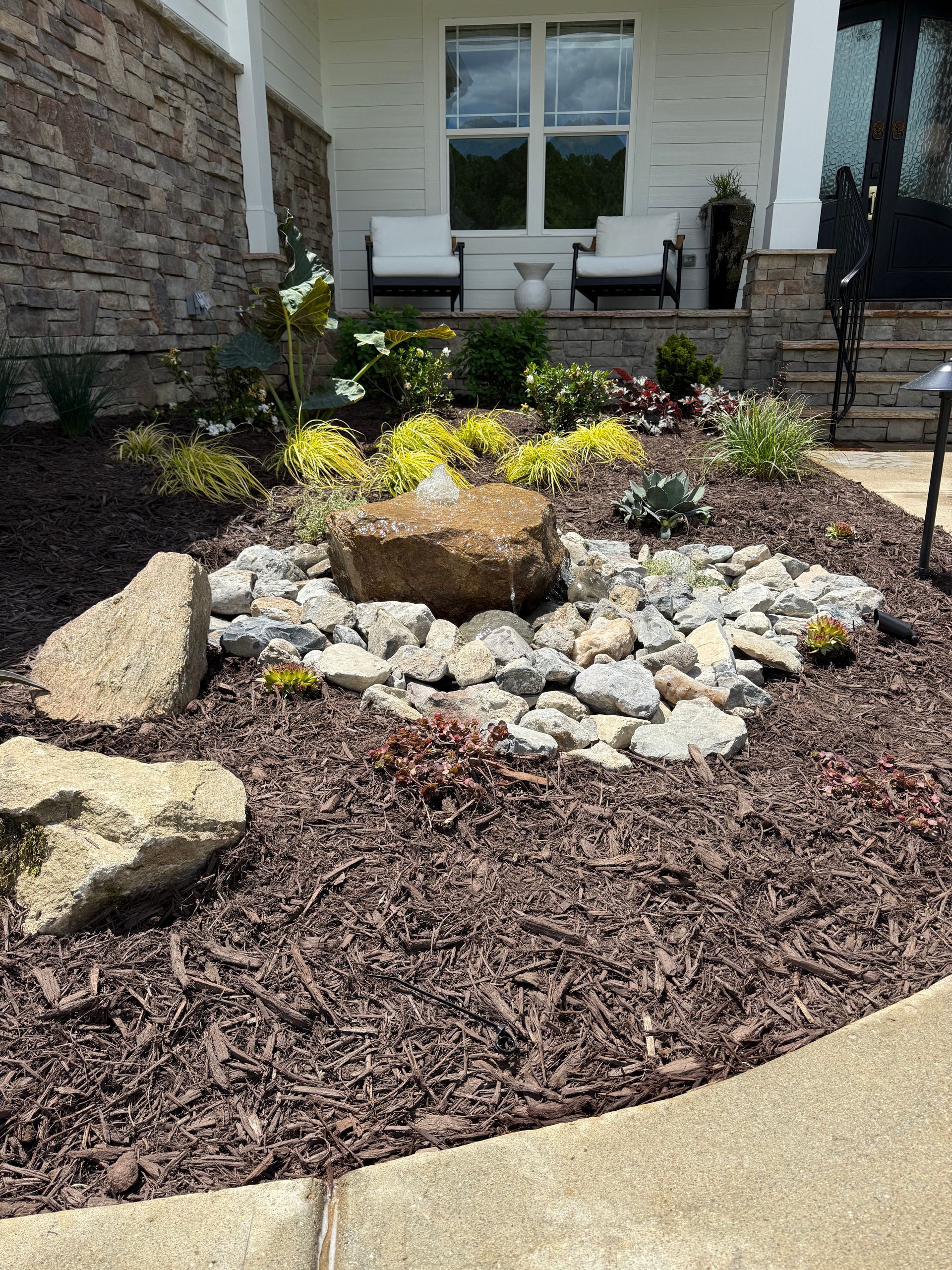 A small rock fountain in a mulch garden bed, surrounded by various plants and rocks, in front of a house.