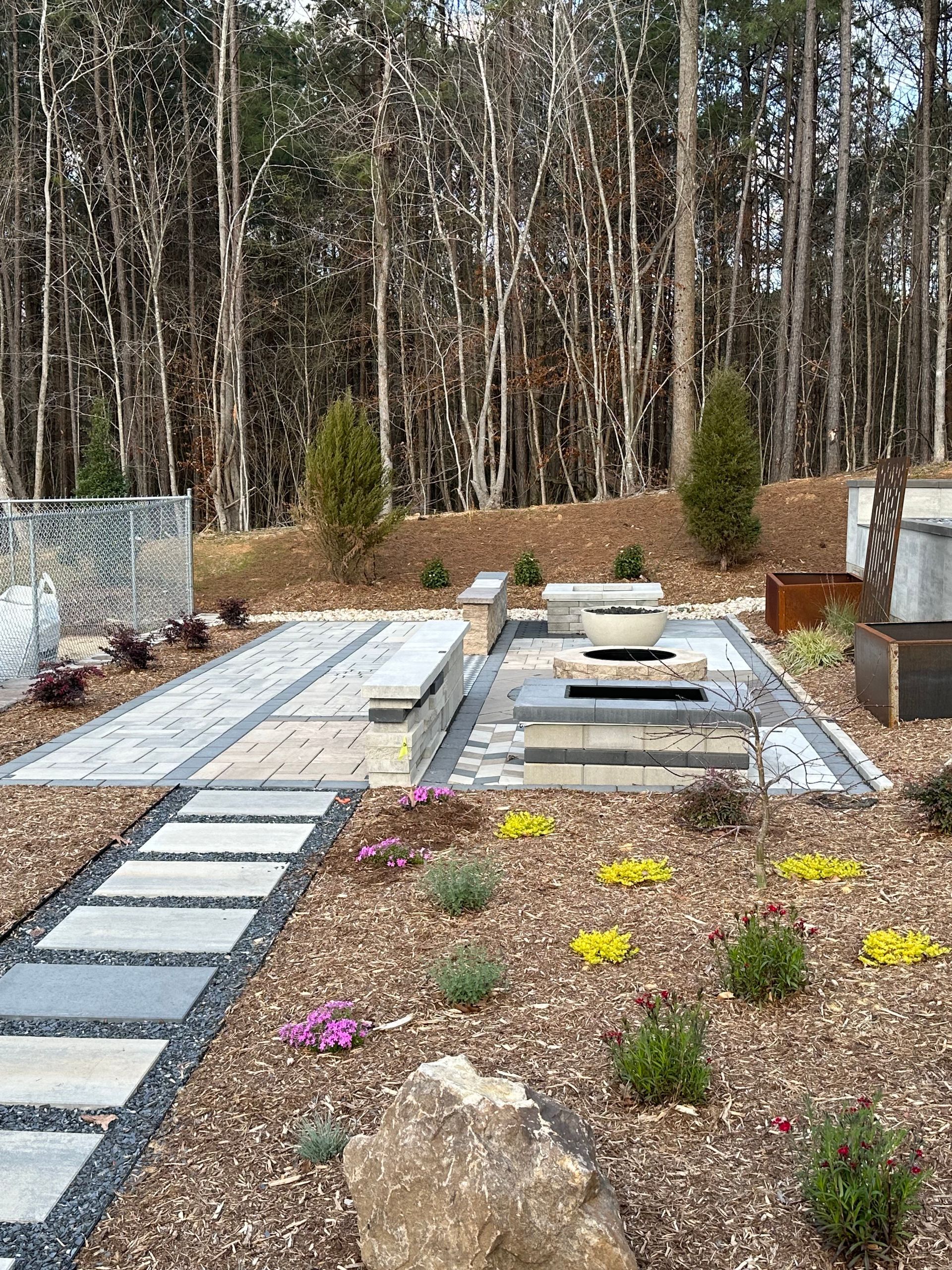 Patio with built-in fire pit, seating, and landscaping; backdrop of trees.