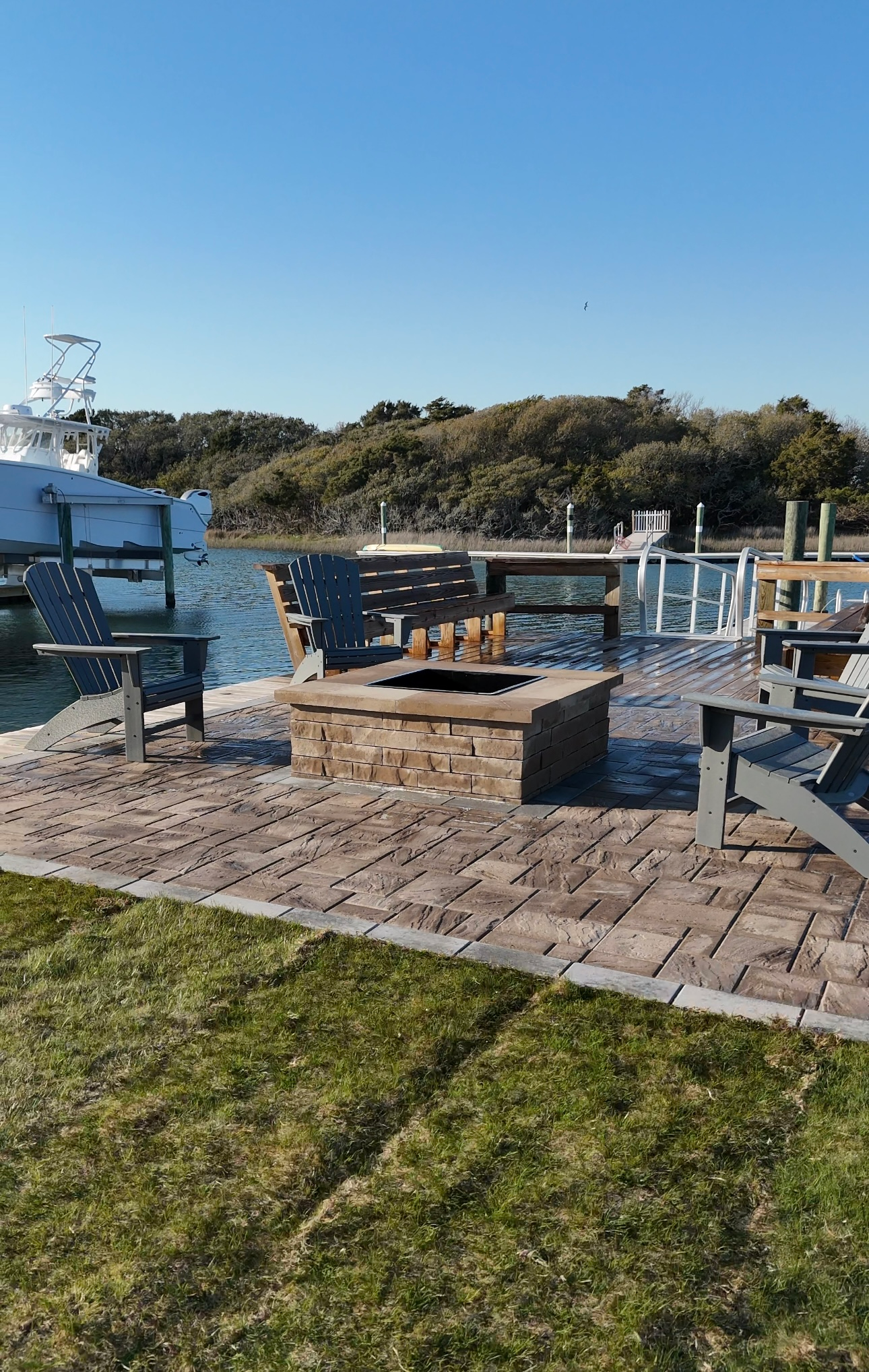 Patio with fire pit, chairs, and dock overlooking water and shoreline on a sunny day.
