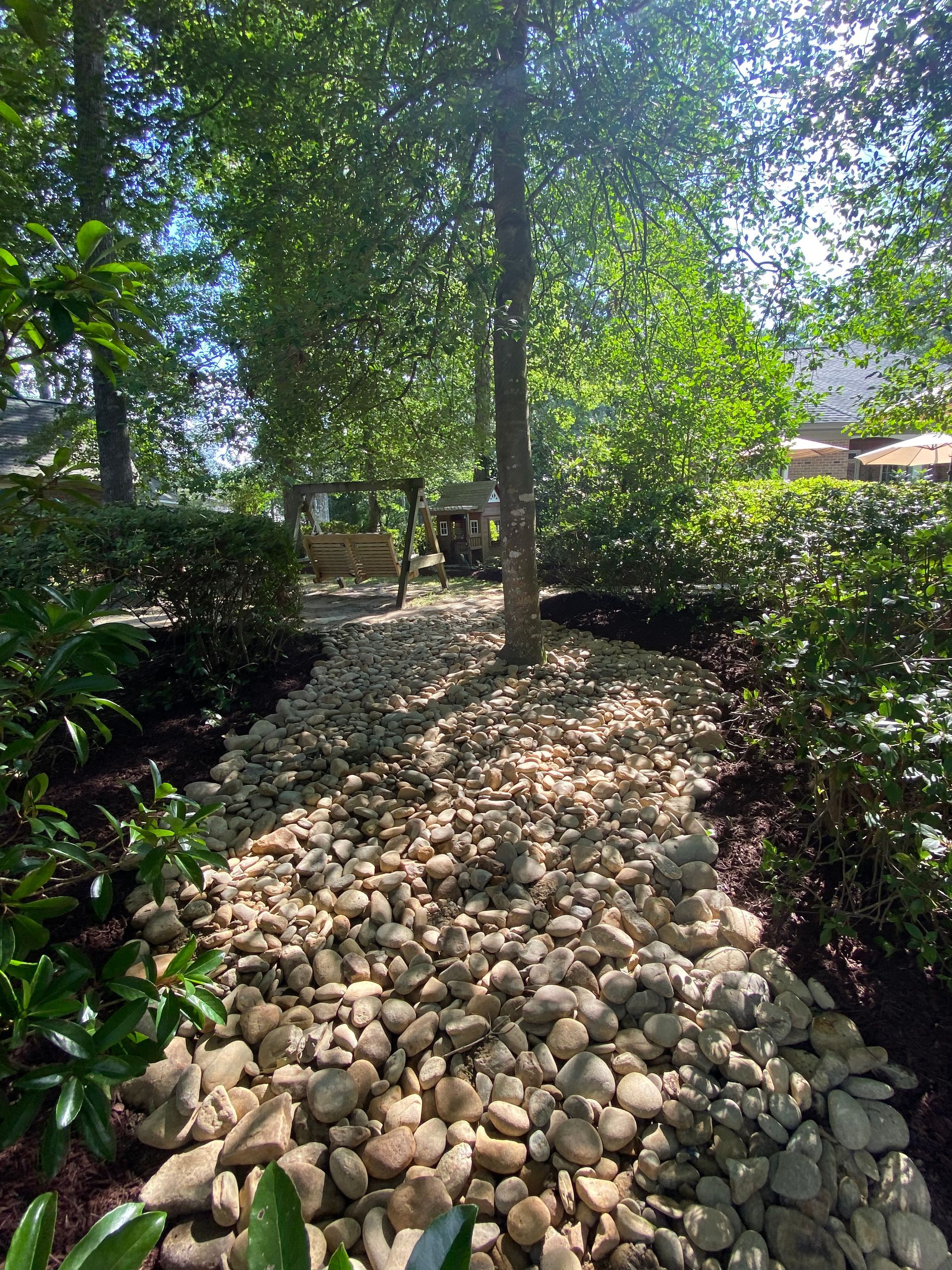 A rock-covered path leads to a wooden swing set under a tree in a lush garden.