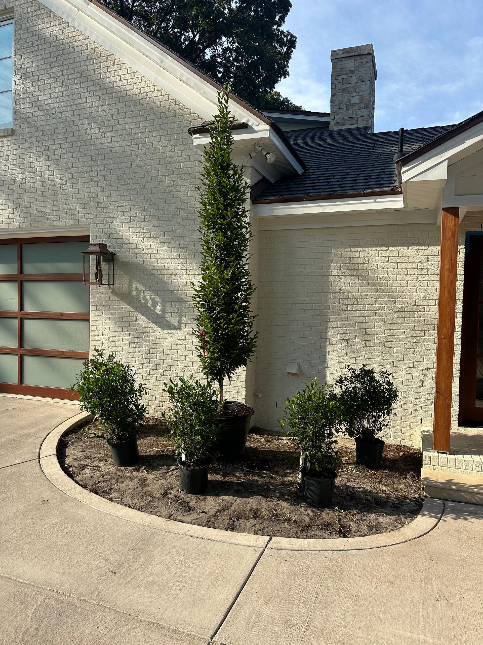 Plants in pots arranged in a bed along the side of a white brick building with a driveway.
