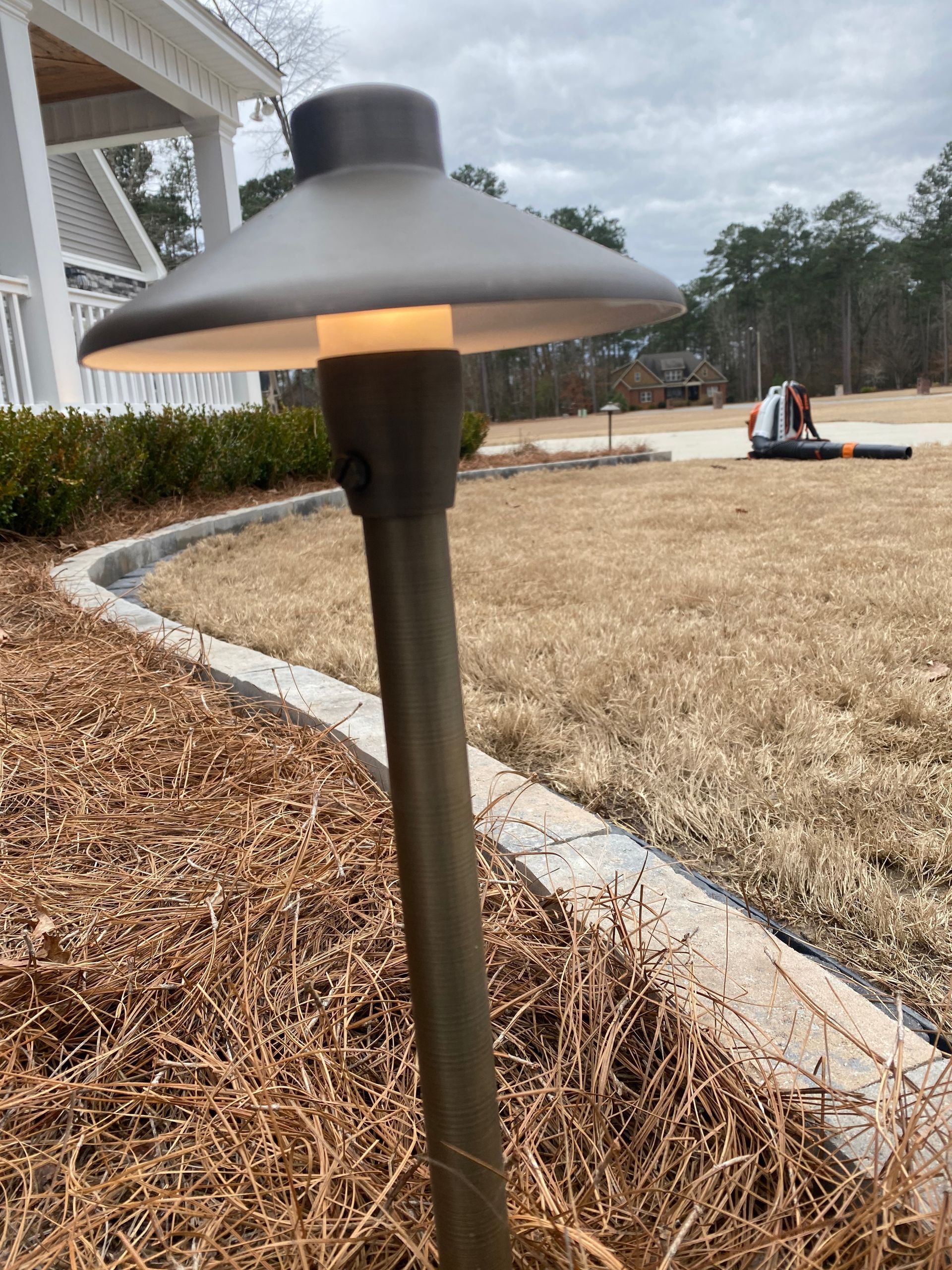 Bronze outdoor pathway light, emitting warm glow, in a grassy area with a house in the background.