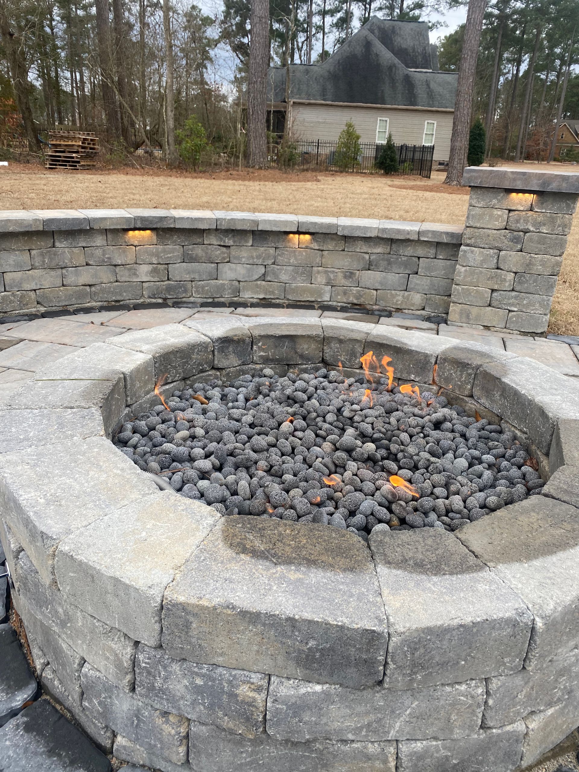 Stone fire pit with flames, surrounded by stone wall and patio in a backyard setting.