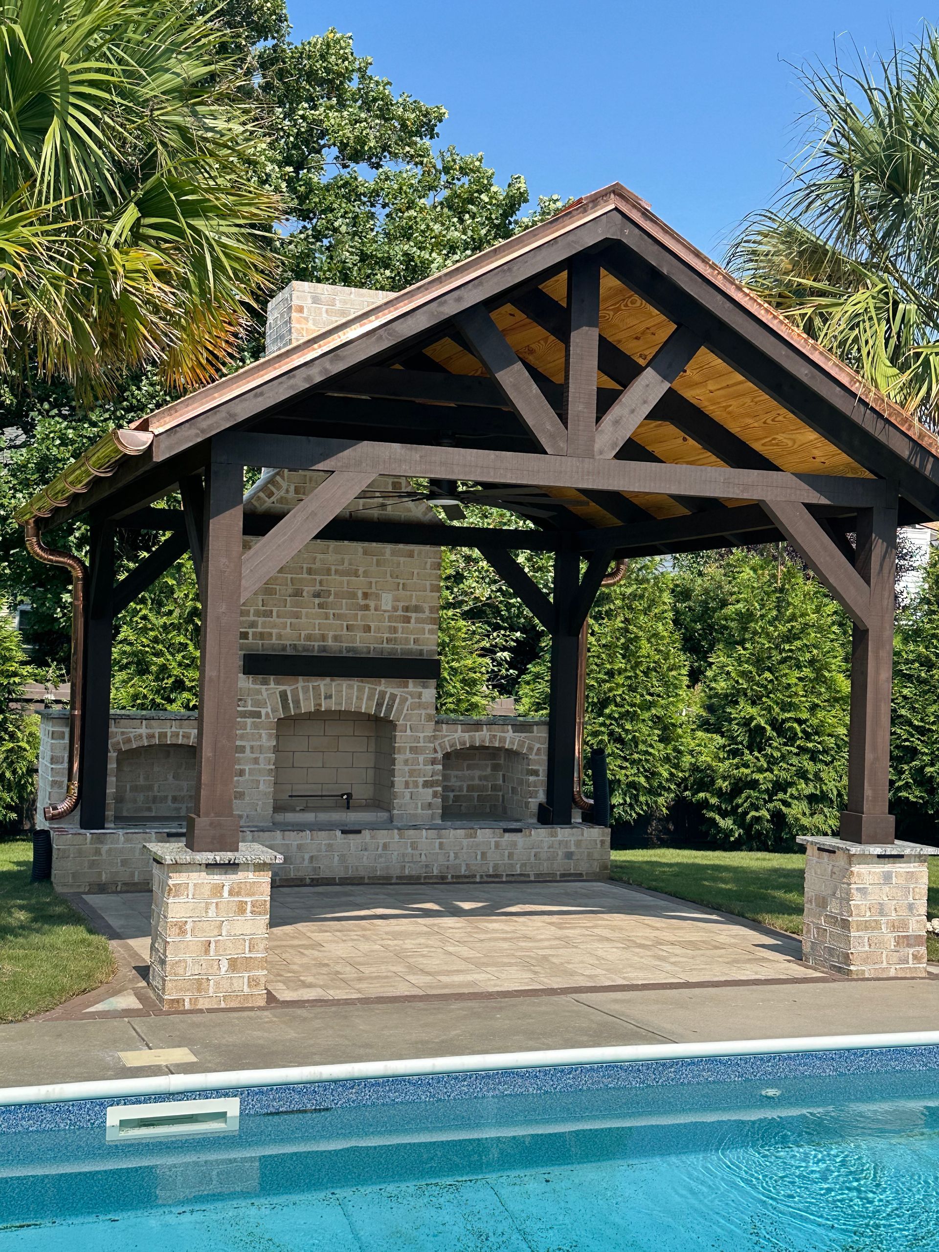 Poolside pavilion with brick fireplace, brown wood frame, and tan roof.