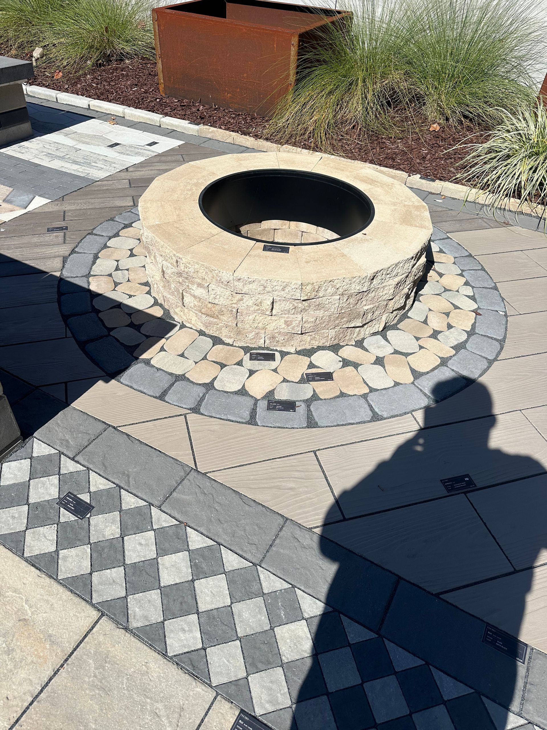Circular stone fire pit on patterned patio, surrounded by pebbles. Sunny outdoor setting.