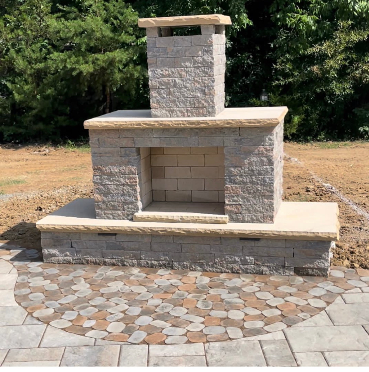 Outdoor stone fireplace with chimney, built on a patio with a circular pattern of pavers.