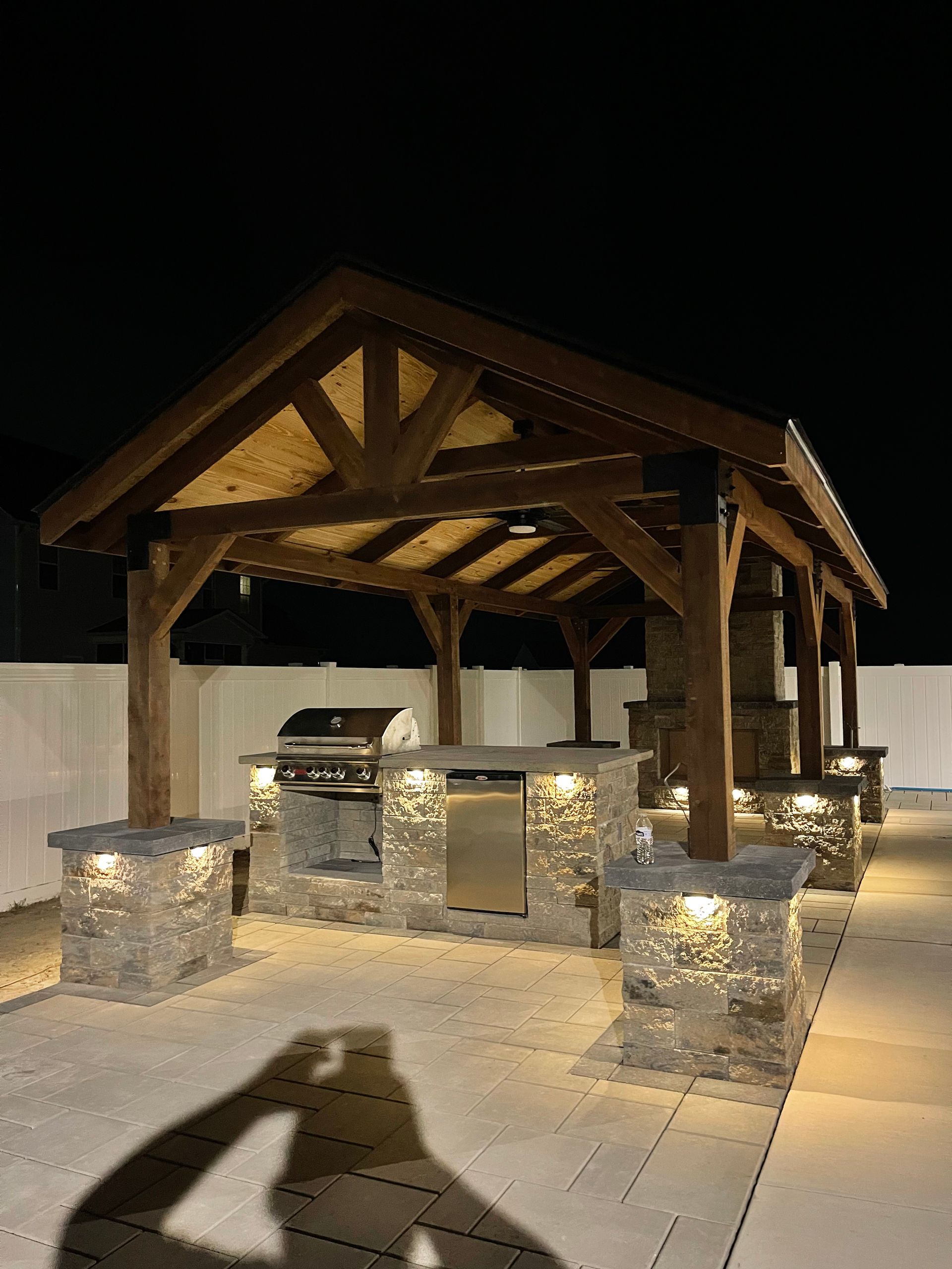 Outdoor kitchen with stone counters, grill, and wood-beam pergola lit at night.
