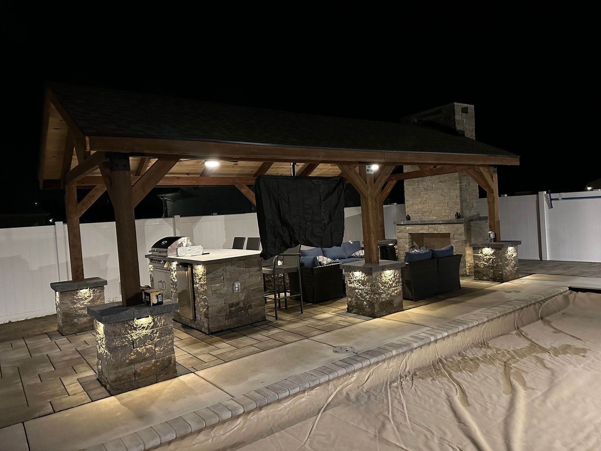 Outdoor patio with stone columns, fireplace, built-in grill, and a covered wooden structure, at night.