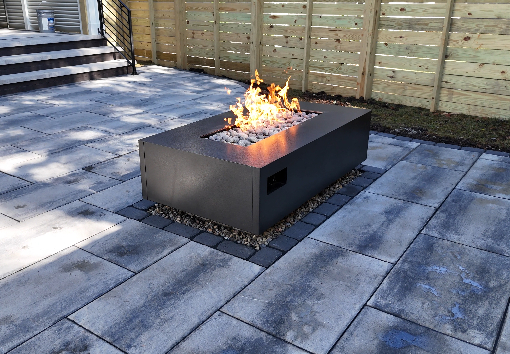 Rectangular fire pit with flames on a gray stone patio, near a wooden fence.
