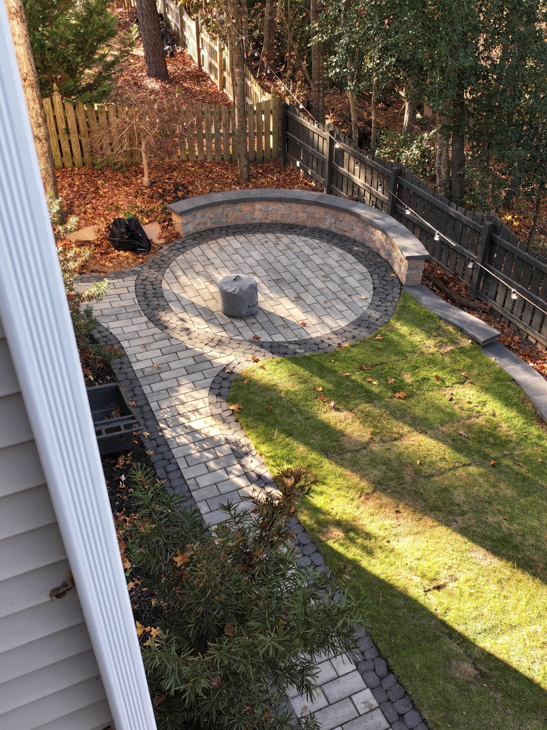 Brick patio and circular seating area in a backyard, with a grassy lawn, fences, and trees.