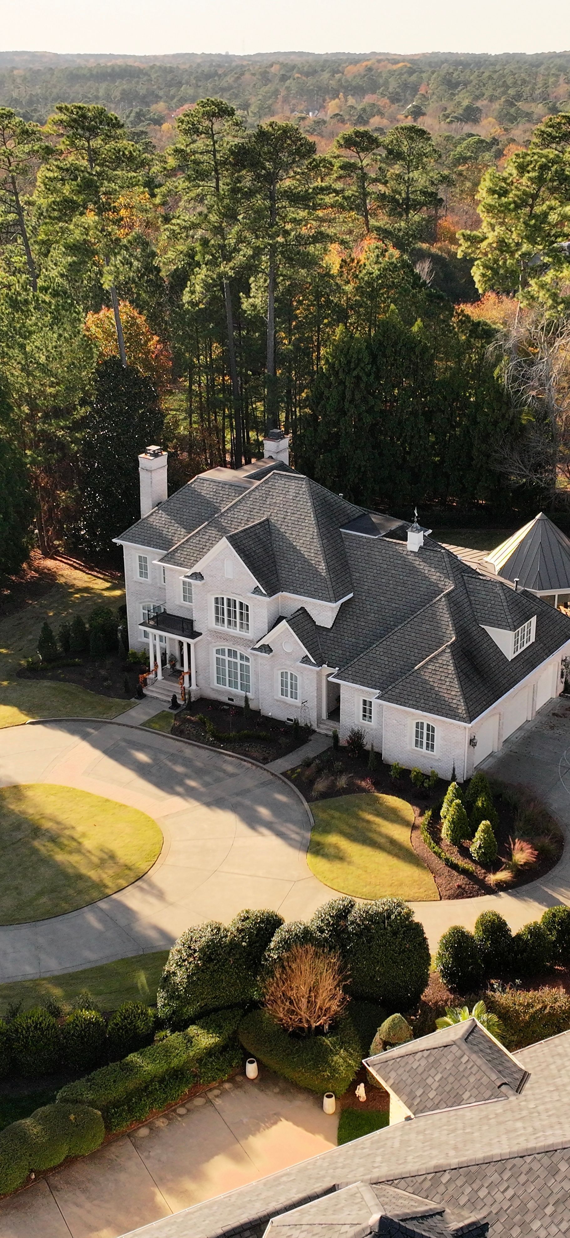 An aerial view of a large white house with a gray roof, surrounded by trees and a driveway.