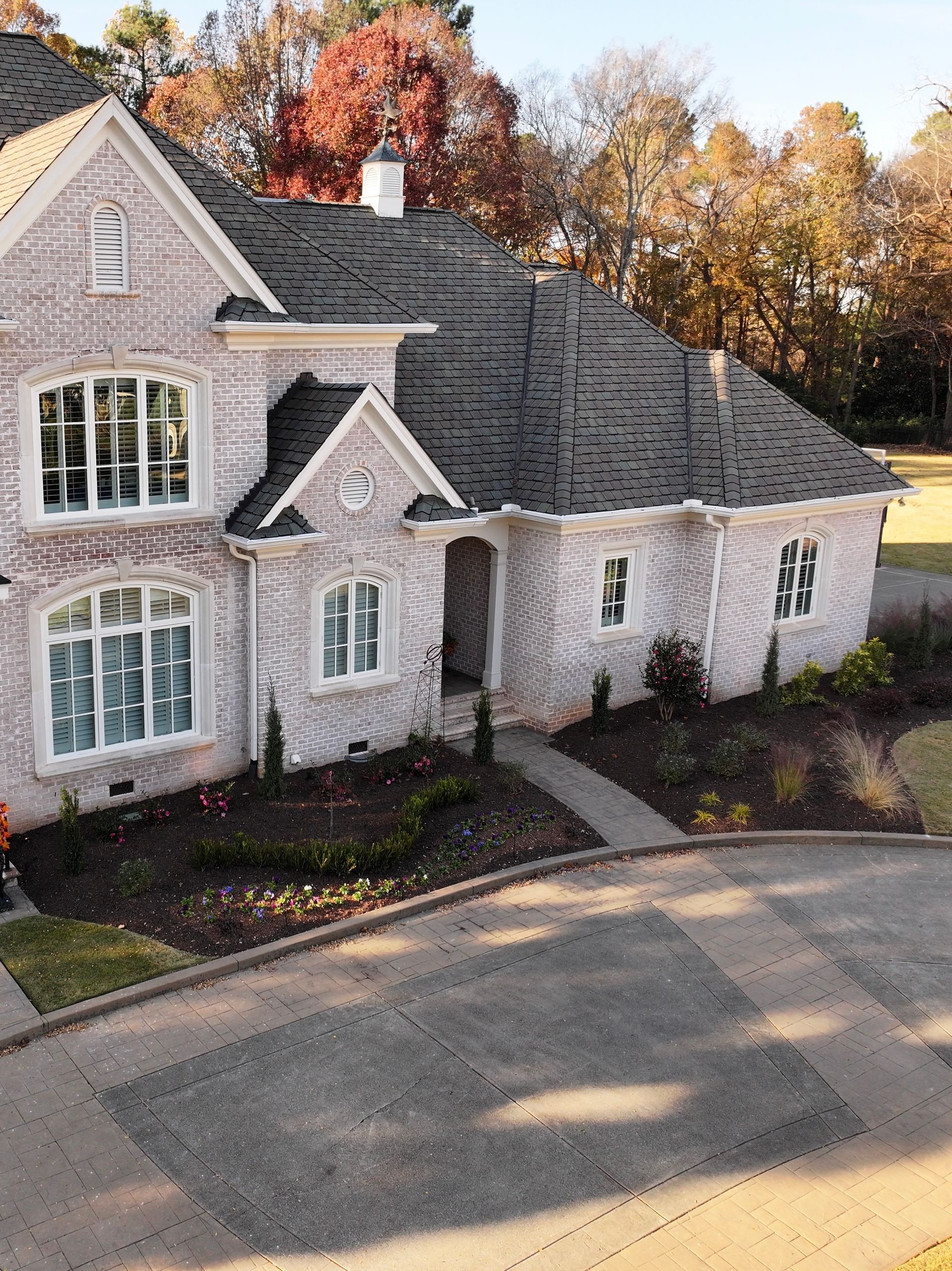 Light brick house with gray roof, multiple windows, and landscaped yard.