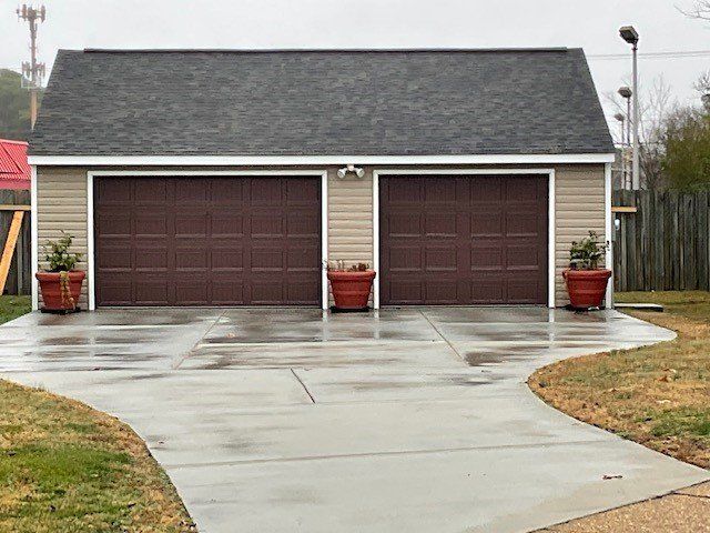 A detached two-car garage with tan siding, a dark roof, brown garage doors, and three large red planters in front.