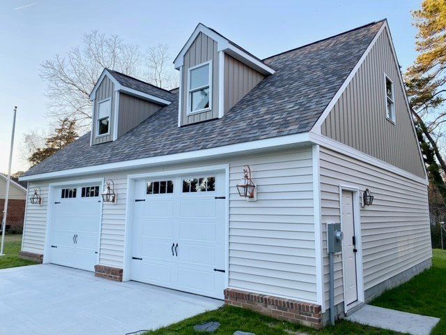 A two-car garage with light siding, a shingled roof, two dormer windows, and a side entry door set against a clear sky.