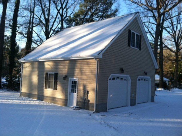 A tan, two-story detached garage with two white bay doors, surrounded by snow and bare trees in late afternoon light.
