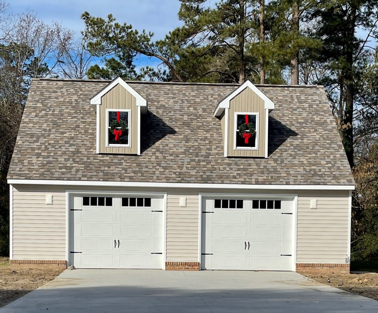 A beige two-car garage with white doors, a grey shingled roof, and two dormer windows decorated for Christmas.