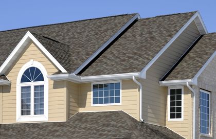 Man Fixing the Roof—General Contractor in Newport News, VA A tan house with brown asphalt shingle roofing, white trim, and multiple rectangular and arched windows against a blue sky.