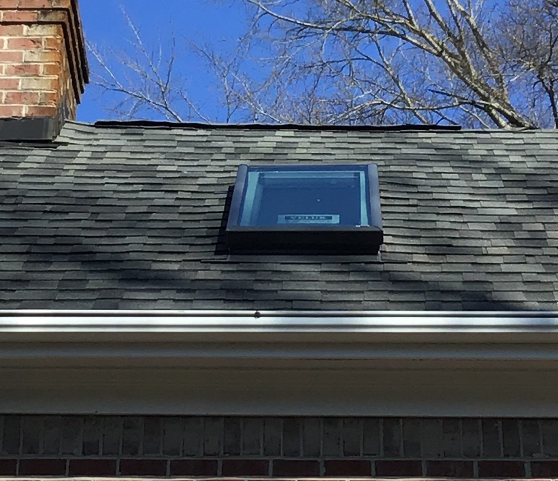 A rectangular skylight installed on a dark gray shingled roof next to a brick chimney under a clear blue sky.