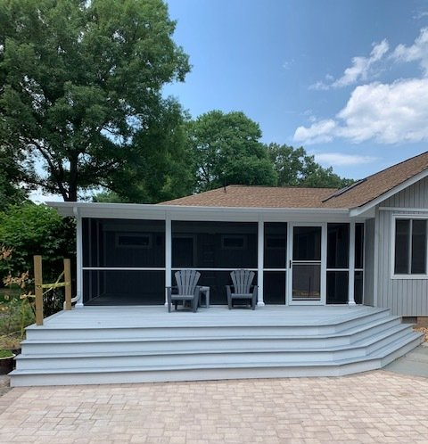 Backyard Deck — Custom Decks in Newport News, VA A grey-painted wooden deck with wide steps and two chairs, attached to a house with a large screened-in porch.