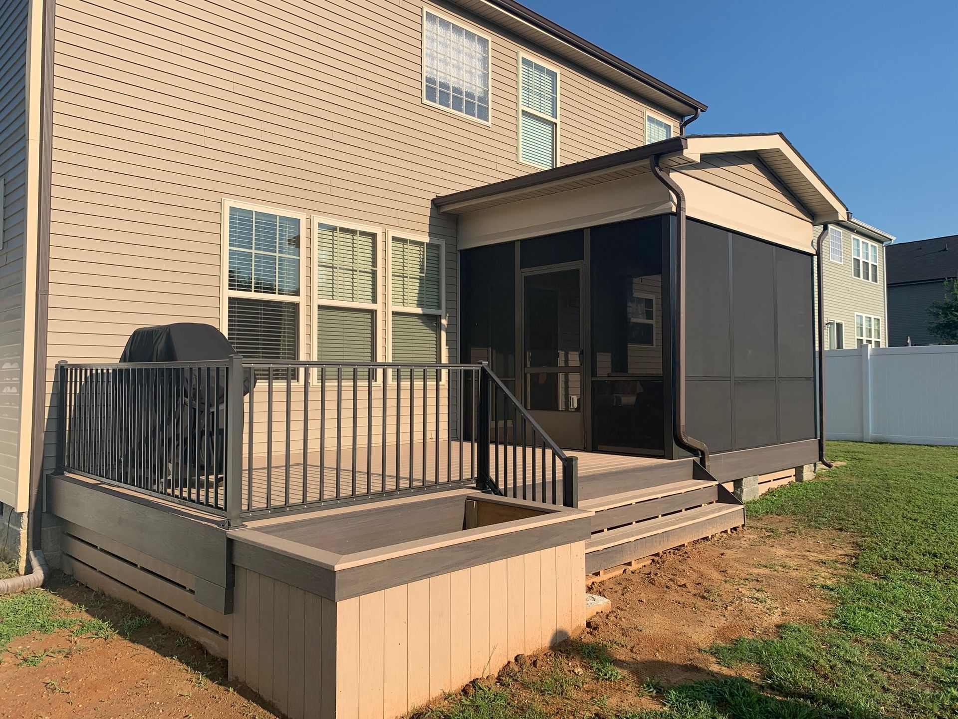 A beige house with a tan composite deck, black metal railings, a covered screened-in porch, and a partially graveled yard.