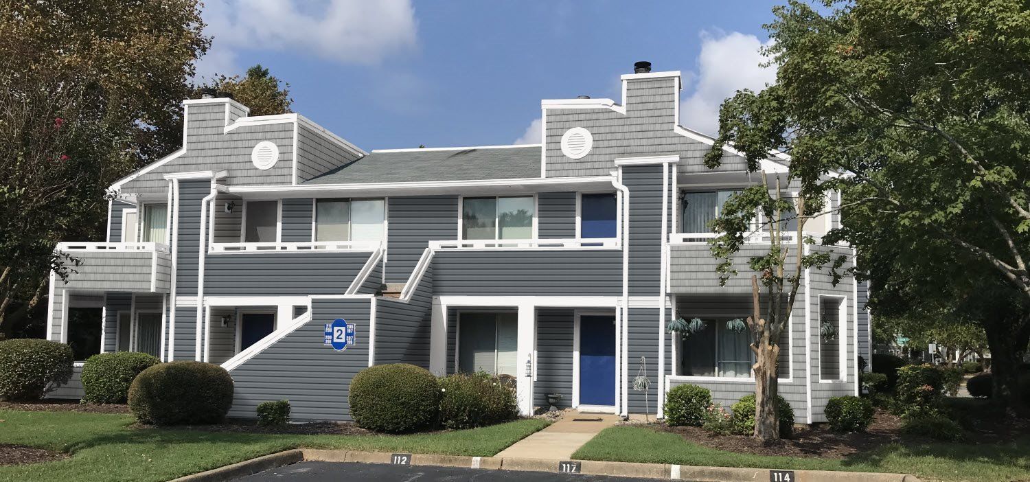 House With Brown Siding — Siding in Newport News, VA A two-story apartment building with gray and white siding, blue doors, and a parking lot in front.