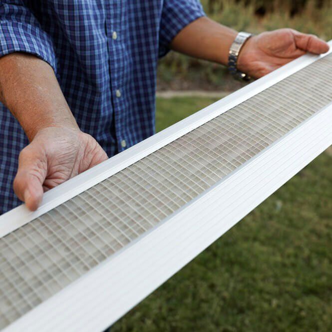 A person holds a long, white gutter guard with a fine mesh screen outdoors.