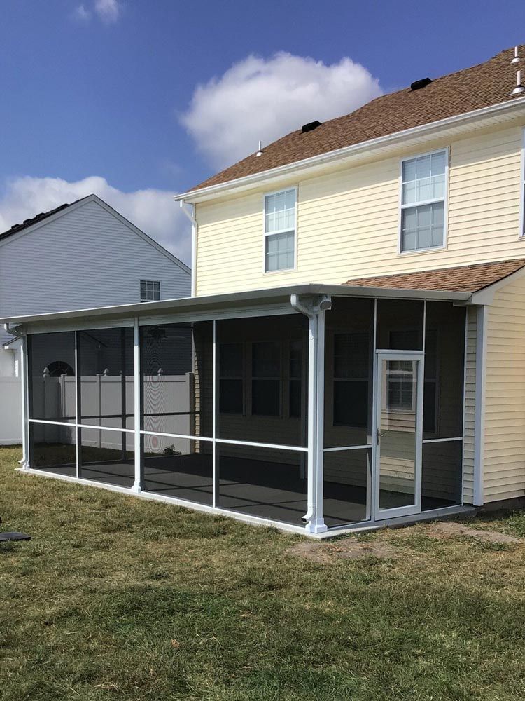A white screened-in patio attached to the back of a two-story, yellow-sided house on a sunny day.