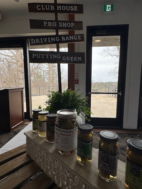 Signage indicating golf amenities above jars of pickles on a counter, with an outside view.