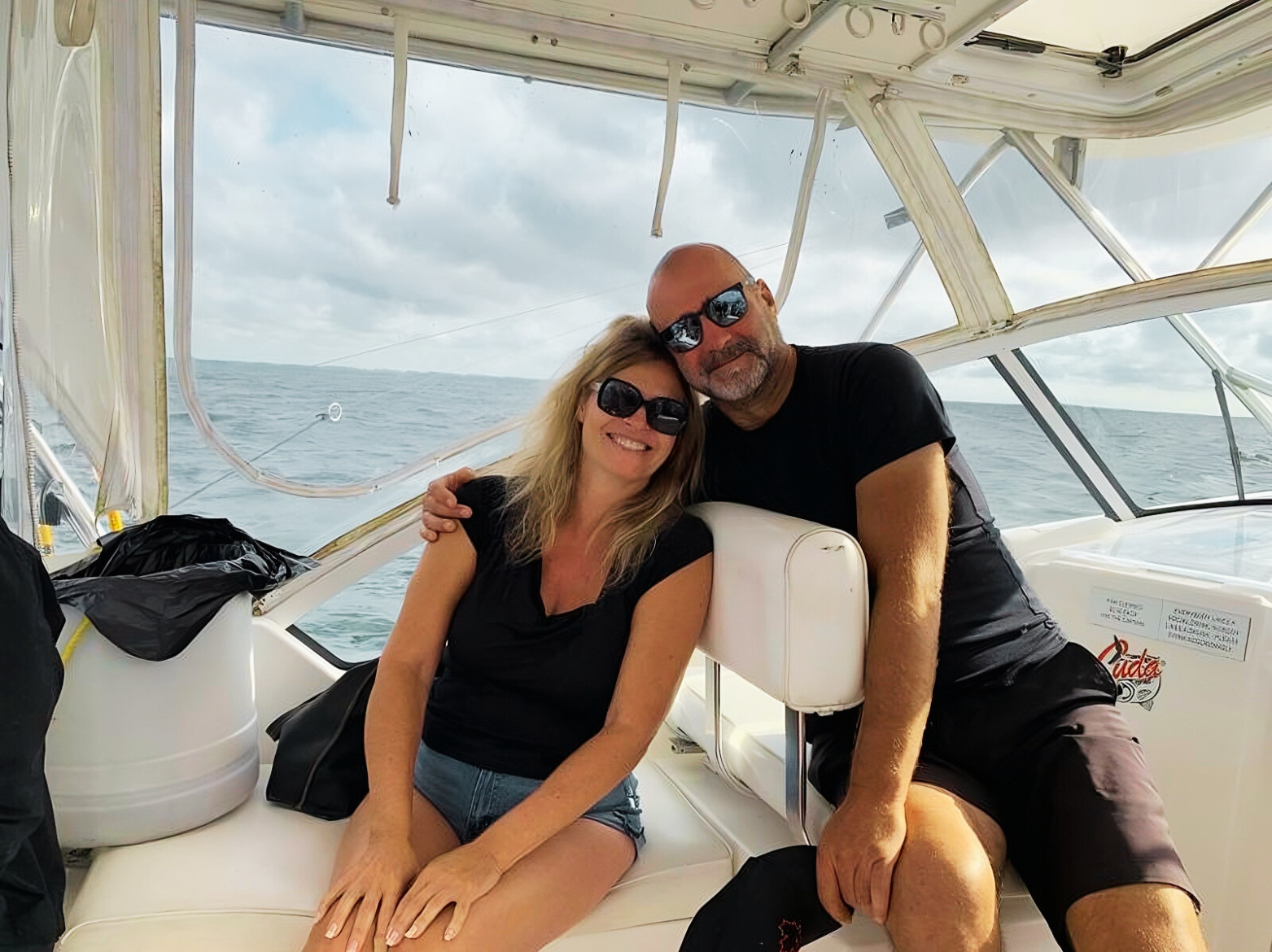 Couple on a boat; woman with blonde hair and man in sunglasses, smiling, posing together. Ocean background.