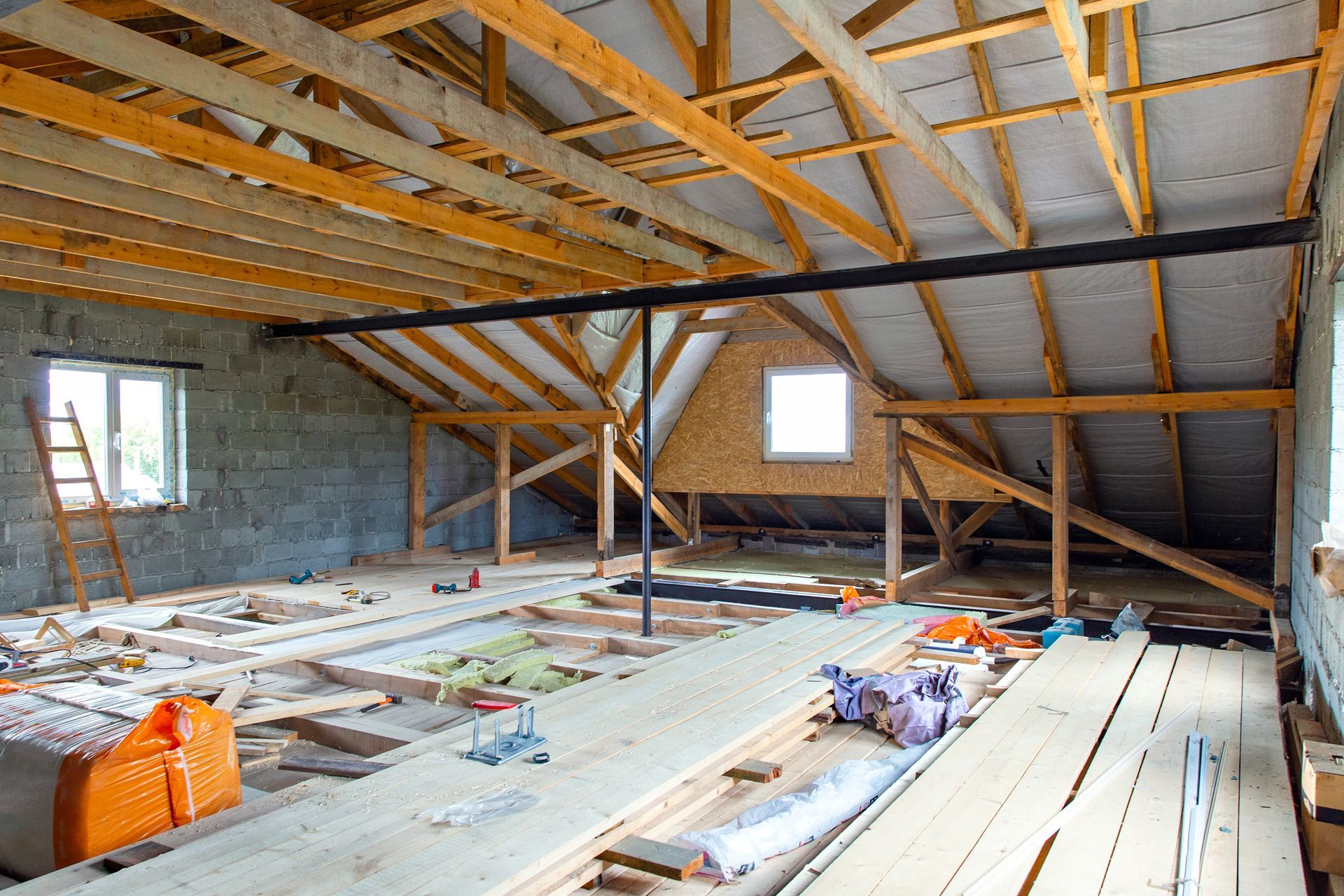 Intérieur d'un toit en construction : poutres en bois, contreplaqué, isolation apparente, fenêtres et outils.